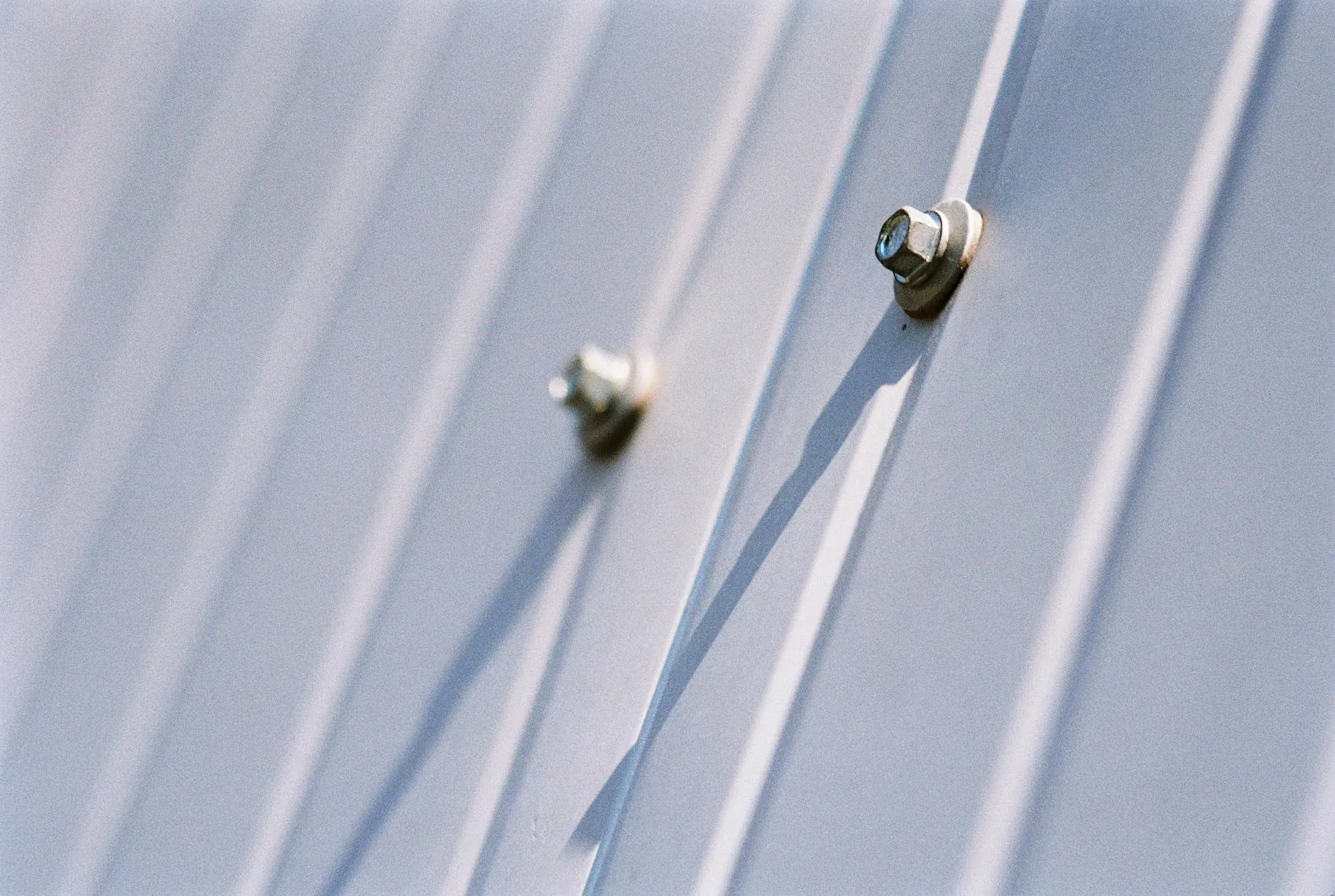 Metal bolt with blue-tinted head fastening corrugated galvanized steel panels together, casting shadow on ridged surface.